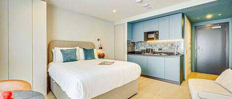 A modern hotel room with a bed, a small table, a sofa, and a kitchenette.