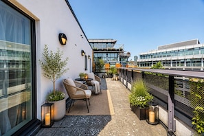 Una terraza en la azotea con mobiliario exterior, plantas en macetas y edificios modernos al fondo.