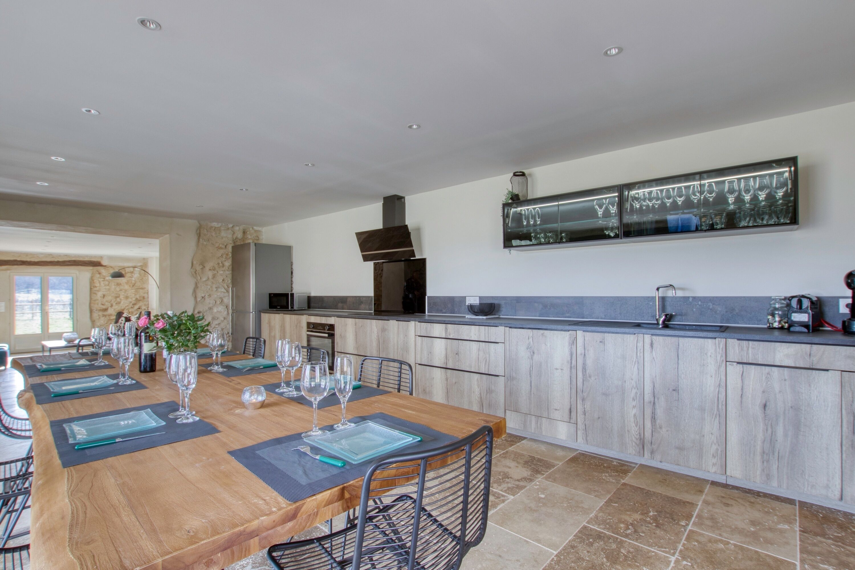 A spacious dining area with a long wooden table set for a meal, flanked by kitchen cabinets and a modern backsplash.