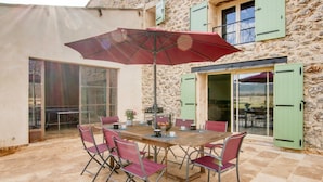 En udendørs terrasse med et træbord og stole, en rød parasol og en stenvæg.