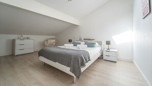 Un dormitorio con tragaluces, una cama con manta gris, dos mesitas de noche y una silla.