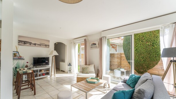 Una sala de estar moderna con un sofá, una mesa de centro y un televisor. Hay una zona de comedor con una mesa y sillas. La habitación tiene ventanas grandes y vistas a la vegetación exterior.