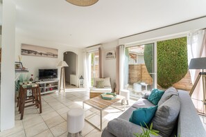 A modern living room with a sofa, coffee table, and a TV. There is a dining area with a table and chairs. The room has large windows and a view of greenery outside. - La Perlière - Avec piscine proche du golf (La Teste-de-Buch)