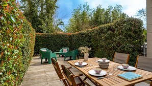 Un patio al aire libre con una mesa de madera preparada para comer, rodeada de sillas verdes y un seto en un lateral.