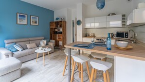 Una sala moderna con un sofá, una mesa de centro y taburetes de bar. Se ve una cocina con gabinetes blancos y una lámpara colgante azul.
