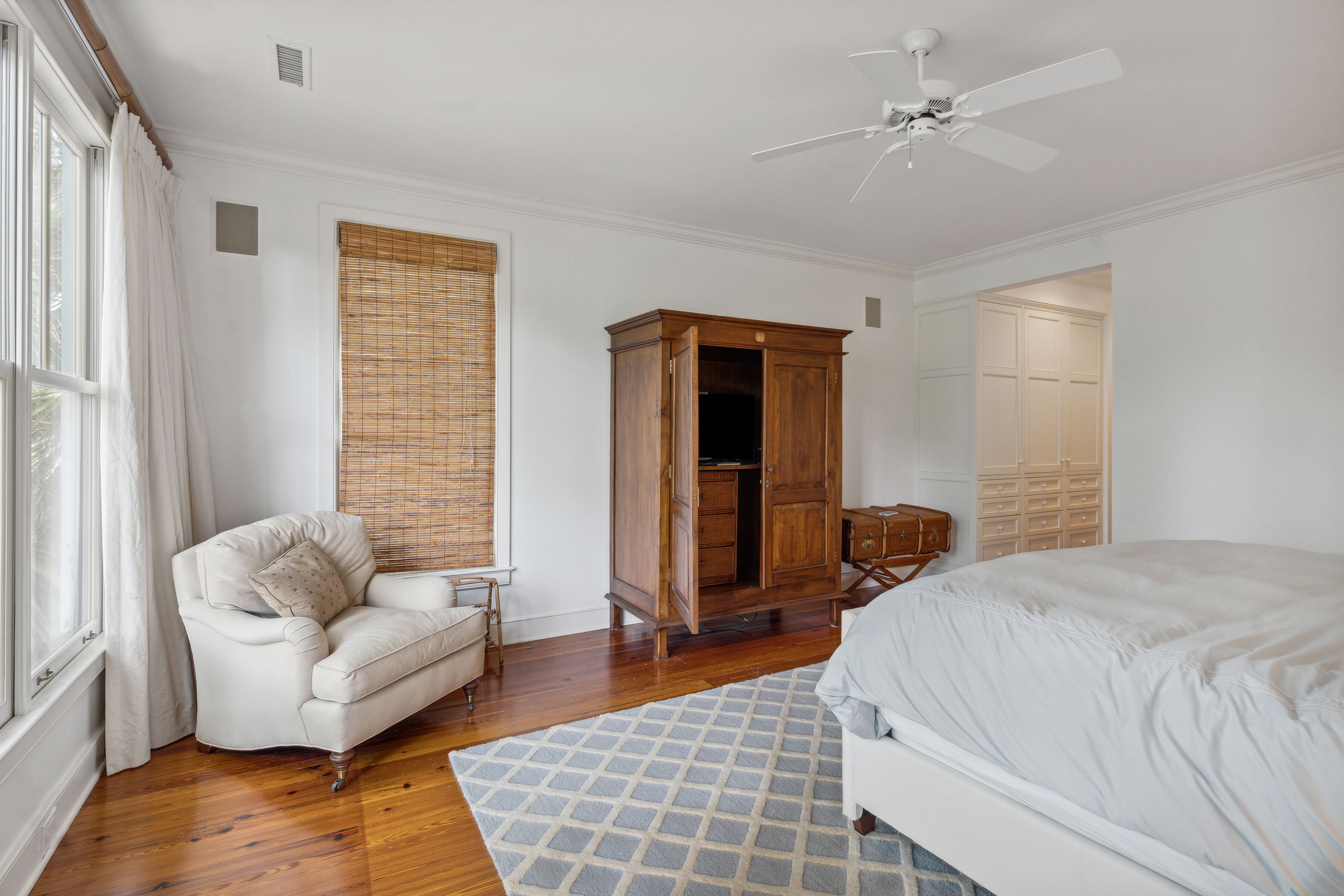 A bedroom with a bed, a wooden wardrobe, a white armchair, and a window with curtains.