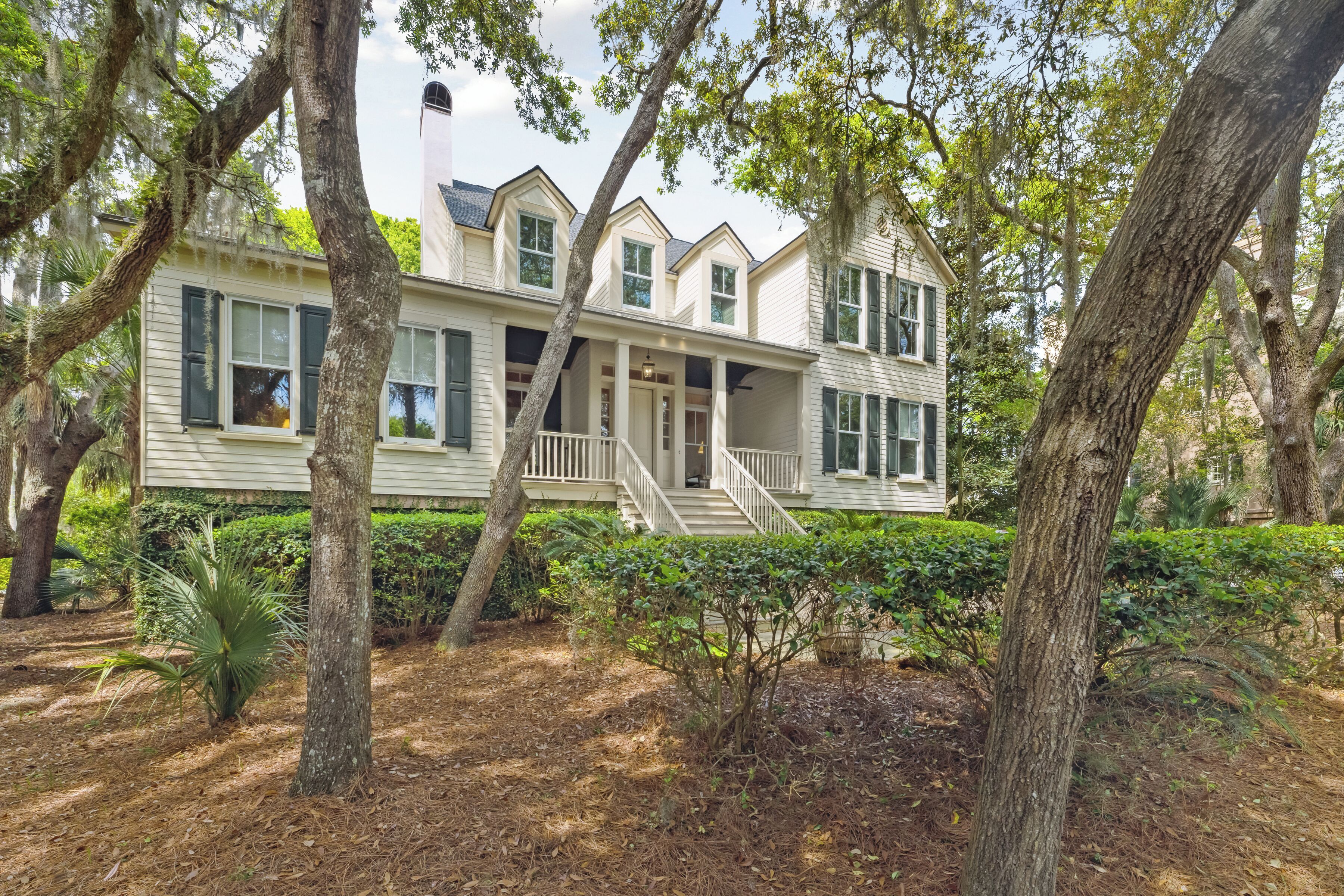 A large, two-story house with a porch and multiple windows, surrounded by trees and shrubs.