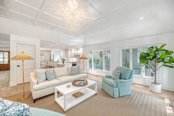 A spacious living room with a white sofa, a blue armchair, and a coffee table. There is a large window with a view of the outdoors.