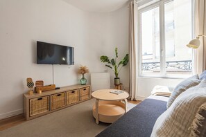 Una habitación de hotel moderna con una televisión de pantalla plana, una mesita consola de madera con cestas, una mesa redonda de madera, un sofá, una planta en maceta y una ventana con vistas a la ciudad.
