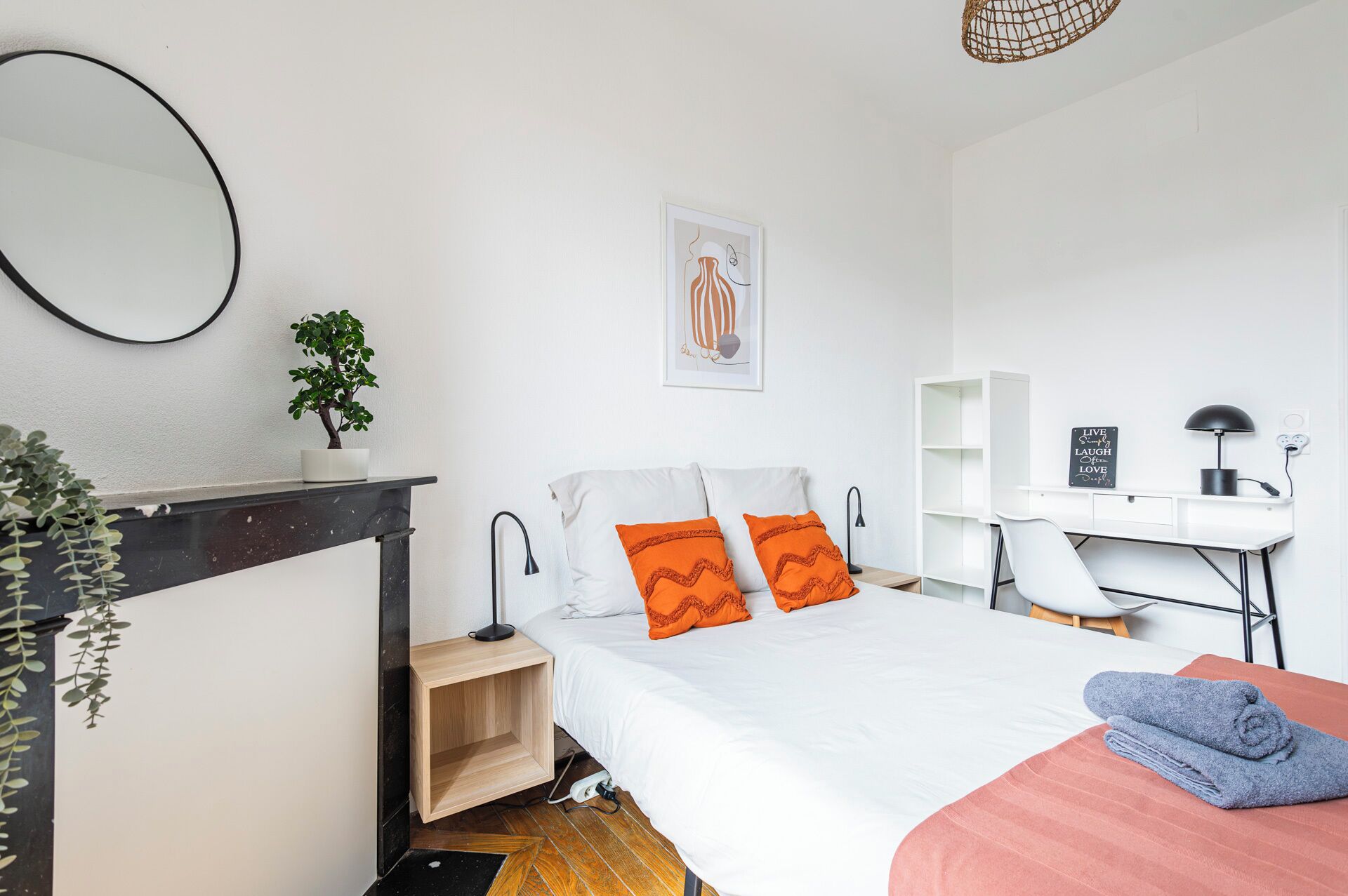Une chambre moderne avec un lit, un bureau et une chaise. On peut voir une petite plante sur la table de chevet et un tableau encadré au mur.