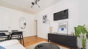 Una sala de estar moderna con una mesa de centro redonda negra, un sofá, una mesa de comedor con sillas, un aparador de madera oscura, un espejo grande, un televisor montado en la pared y una planta en maceta.