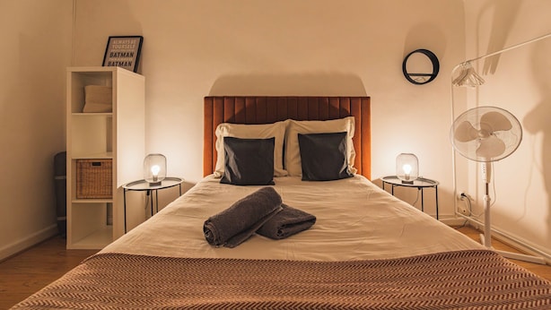 Una cama bien hecha con colcha marrón de estampado romboidal, dos almohadas y una toalla doblada sobre un colchón blanco. Hay dos mesitas de noche con lámparas, un espejo circular montado en la pared y un ventilador de pie. A la izquierda, una estantería blanca con cestas.