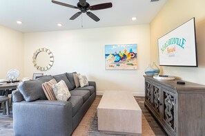 Un salon moderne avec un canapé gris, une table basse, un ventilateur de plafond, un téléviseur fixé au mur diffusant une image du « Margaritaville Resort Orlando », et un tableau sur le thème de la plage.