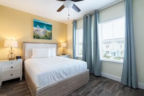 A bedroom with a bed, nightstands, a ceiling fan, a painting, and a window with curtains. - Margaritaville Townhome Retreat (Kissimmee)