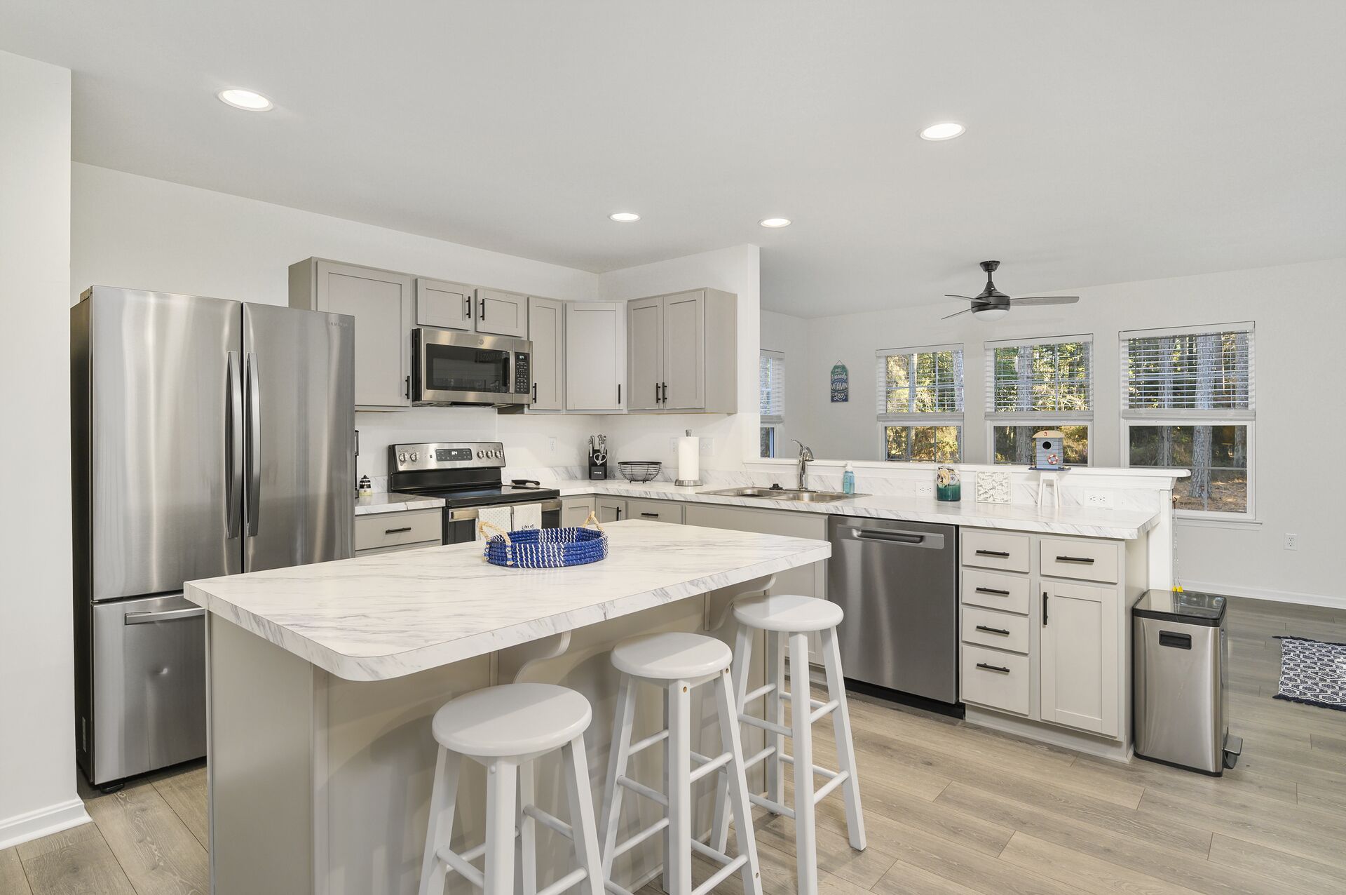 A modern kitchen with white cabinets, stainless steel appliances, a marble island with stools, and a view of the outdoors through large windows.