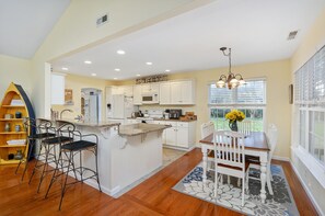 A kitchen with white cabinets, a central island, and a dining area with a table and chairs. - The Sweets Spot by Seaside Vacations & Sales (Greenbackville)