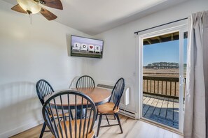 A dining area with a wooden table and four chairs, a ceiling fan, and a television displaying 'LOVE' on the wall. - Assateague Inn 102 by Seaside Vacations & Sales (Chincoteague)