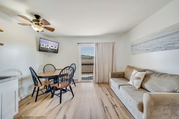 A room with a dining table, chairs, a sofa, a ceiling fan, a TV, and a view through a window. - Assateague Inn 102 by Seaside Vacations & Sales (Chincoteague)
