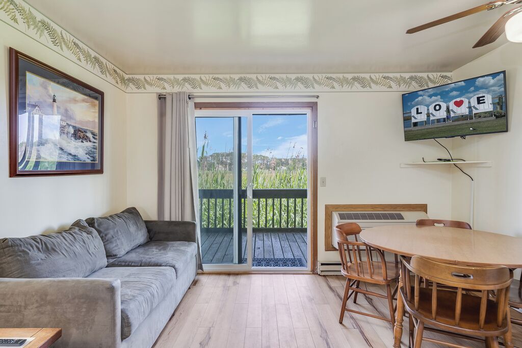A living room with a sofa, a dining table with chairs, a TV mounted on the wall, and a sliding glass door leading to a balcony.