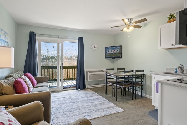 A cozy living room with a sofa, a dining table with chairs, a TV mounted on the wall, and a ceiling fan. - Assateague Inn 105 by Seaside Vacations & Sales (Chincoteague Island)