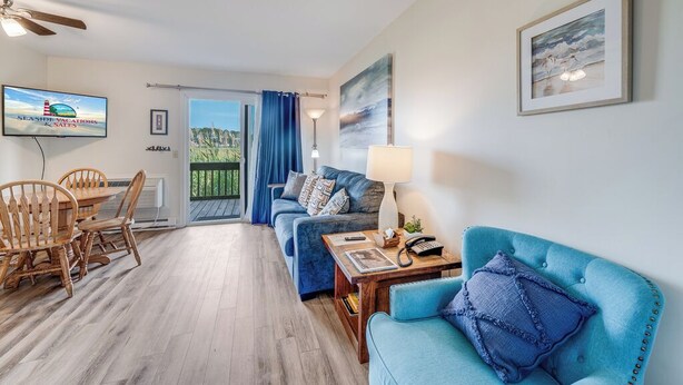 Un salon avec un canapé bleu, une table à manger en bois avec des chaises, un téléviseur et un balcon agrémenté de rideaux bleus.
