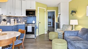 Eine moderne Küche mit weißen Schränken, einer blauen Fliesenwand und einem Edelstahlbackofen. Ein Essbereich mit Holztisch und Stühlen sowie ein Wohnzimmer mit blauem Sofa und Poufs.