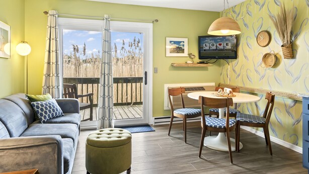 Ein Wohnzimmer mit einer Couch, einem Hocker und einem Esstisch mit Stühlen. An der Wand befindet sich ein Fernseher und ein Fenster mit Ausblick.