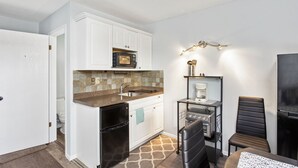 A compact kitchen with white cabinetry, a microwave, a coffee maker, and a small dining area with a chair and table. - Assateague Inn 111 by Seaside Vacations & Sales (Chincoteague Island)