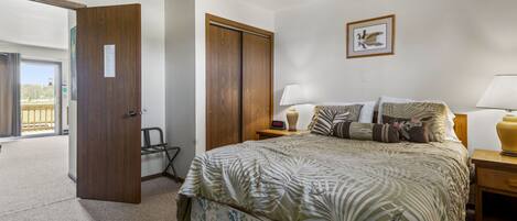 Un dormitorio con una cama, dos mesitas de noche, un armario de madera, una puerta que da a un balcón y un cuadro enmarcado en la pared.