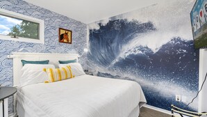 A bedroom with a large wave mural on the wall, a bed with white bedding and colorful pillows, and a framed picture above the bed. - Assateague Inn 213 by Seaside Vacations & Sales (Chincoteague Island)