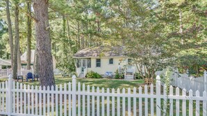 A single-story house with a front porch, surrounded by a white picket fence and trees. - Teague Time by Seaside Vacations & Sales (Chincoteague)