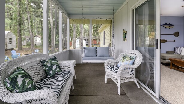 A covered porch with wicker furniture, a sliding glass door, and a view of the surrounding trees. - Teague Time by Seaside Vacations & Sales (Chincoteague)