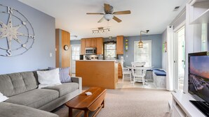 A modern living room with a sofa, coffee table, and a TV. A kitchen with wooden cabinets and a dining area is visible in the background. - Teague Time by Seaside Vacations & Sales (Chincoteague)