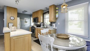 A modern kitchen with wooden cabinets, a microwave, a sink, and a dining area with a glass table and white chairs. - Teague Time by Seaside Vacations & Sales (Chincoteague)