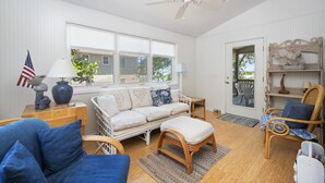 A spacious living room with a white sofa, blue armchairs, and a wooden coffee table. A ceiling fan and a large window provide natural light. - The Happy Crab by Seaside Vacations & Sales (Chincoteague Island)
