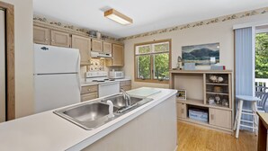 A compact kitchen with a refrigerator, stove, microwave, and a sink. There is a window with blinds, a painting on the wall, and a small table with a chair. - The Happy Crab by Seaside Vacations & Sales (Chincoteague Island)