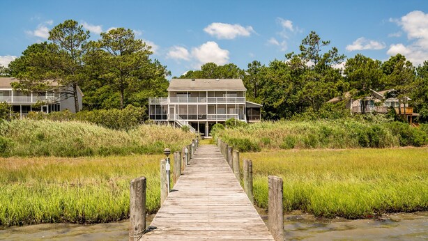 A wooden boardwalk leading to a two-story building with a balcony, surrounded by grassy fields and trees. - The Happy Crab by Seaside Vacations & Sales (Chincoteague Island)