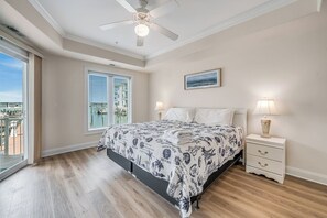 A bedroom with a bed, bedside lamps, a ceiling fan, a window with blinds, and a painting on the wall. - Sunset Bay 110 by Seaside Vacations & Sales (Chincoteague Island)