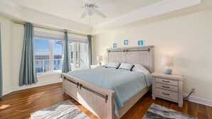 Une chambre à coucher comprenant un lit, des tables de chevet, un ventilateur de plafond, une fenêtre avec des rideaux et une vue sur l’océan.