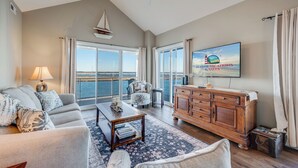 A spacious living room with a large window offering a view of the sea, a comfortable sofa set, a wooden cabinet, and a television displaying a scenic vacation advertisement. - Sunset Bay 319 by Seaside Vacations & Sales (Chincoteague Island)