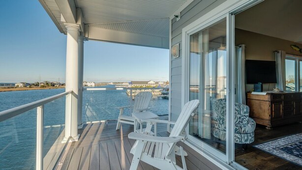 A wooden deck with white Adirondack chairs, a glass door leading to a room, and a view of the water and distant buildings. - Sunset Bay 319 by Seaside Vacations & Sales (Chincoteague Island)