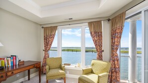 A room with two armchairs, a wooden desk, and a view of a body of water through large windows. - Sunset Bay 319 by Seaside Vacations & Sales (Chincoteague Island)
