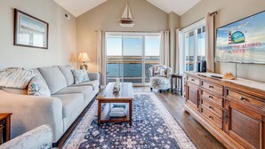 A spacious living room with a large window offering a view of the sea, a comfortable sofa, a coffee table, and a wooden cabinet. - Sunset Bay 319 by Seaside Vacations & Sales (Chincoteague Island)