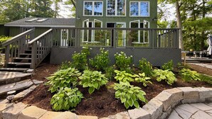 A green wooden house with a porch, a staircase, and a garden with various plants. - Enjoy Beautiful Sunrise & Sunset in Your Private Island Paradise (Duoro-Dummer)