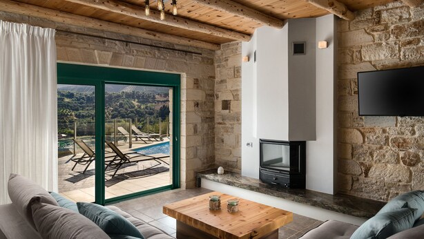 A cozy living room with a stone wall, a wooden ceiling, a fireplace, and a view of a pool and mountains. - Villa Piedra (Platanias)