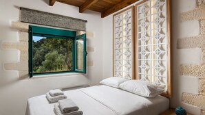 A bedroom with a bed, a window with a view of greenery, and a decorative screen. - Villa Piedra (Platanias)