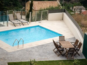 A rectangular swimming pool with clear blue water, surrounded by a tiled deck. There are lounge chairs and a wooden table with chairs on the deck.