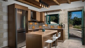 A modern kitchen with a stainless steel refrigerator, wooden cabinets, a bar area with stools, and a view of the outdoors through a window. - Villa Piedra (Platanias)