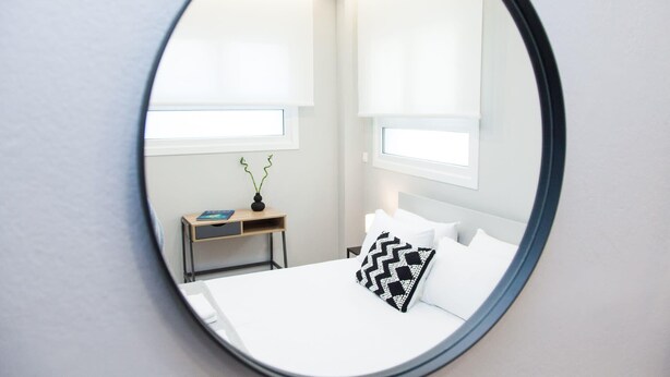 A bedroom with a round mirror, a bed with white bedding, a small bedside table with a book and a vase, and two windows with blinds.