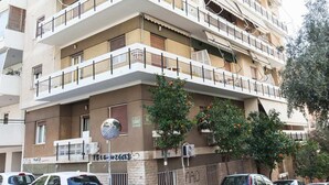 A multi-story residential building with balconies, parked cars, and orange trees.